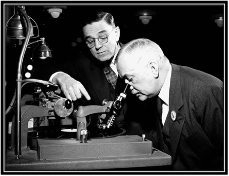 L'honorable Major-général L.R. LaFlèche, ministre des Services nationaux de guerre, examine un disque de phonographe sous les yeux de F.R. Deakins, président de RCA Victor. 